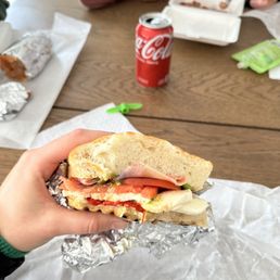 Fats Sandwich - A person holds a sandwich with ham, tomato, lettuce, and cheese, wrapped in foil, over a table with a soda can and other food items.