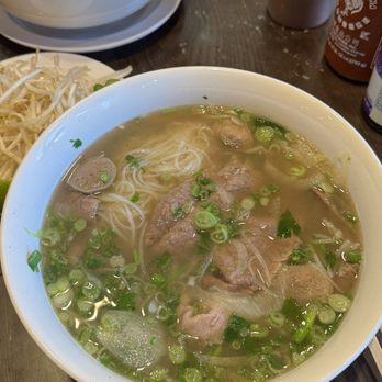Pho Che - Two bowls of Vietnamese pho with beef slices, meatballs, and chopped green onions are on a dark table, accompanied by condiments and utensils. - Vietnamese Near Me - Flushing, New York