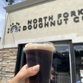 North Fork Doughnut - A person holds a cup of black coffee outside North Fork Doughnut Co, with the business sign visible on the building. - Donuts Near Me - Mattituck, New York