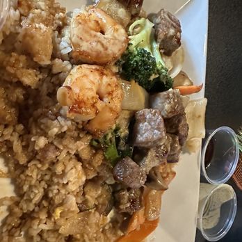 Asian Cravings - A white plate holds a serving of fried rice with a golden-brown color, topped with shrimp. Next to the rice are pieces of beef, broccoli, and mixed vegetables including carrots and onions, all coated in a sauce. Two small clear containers of sauce are placed beside the plate. The presentation suggests a Chinese or Asian-inspired meal. - Thai Near Me - Akron, Ohio