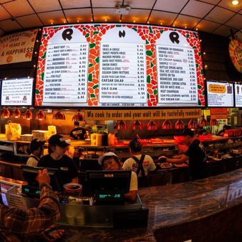 Roll-N-Roaster - A busy fast-food restaurant interior with a curved counter, digital menu boards, and customers placing orders, featuring a colorful menu and warm lighting. - American Near Me - Brooklyn, New York