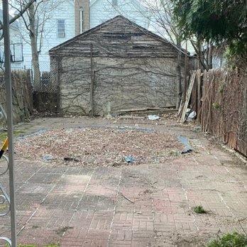 Smart General Contracting - Before. - A backyard scene featuring a brick patio with some uneven and cracked bricks, a small dirt area with sparse vegetation, and a weathered wooden shed at the back. The yard is enclosed by wooden fences on both sides, with trees visible beyond the fences. The shed appears old, with vines or wires growing on its exterior. The image is taken from under a covered porch or awning, with string lights hanging overhead. - Stucco Services Near Me - Brooklyn, New York