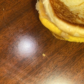 Five Guys - blonde hair ^ - The image shows a breakfast sandwich with egg and possibly sausage or ham, partially visible on a wooden table. There is a small piece of food and a thin hair on the table surface. The focus is on the table's surface and the edge of the sandwich, with the sandwich occupying the upper right corner of the image. - Burgers Near Me - Lubbock, Texas