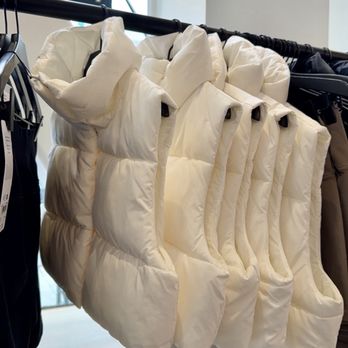 ALO - Down jacket - Several white puffer vests are hanging on a black clothing rack inside a retail store. The vests are arranged side by side, with some showing the front and others the back. A window with a partial view of the outside is visible in the background, allowing natural light into the space. - Men's Clothing Near Me - Pasadena, California