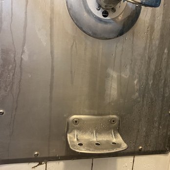 Hays Communities YMCA - Soap scum and black mold. Black mold can be dangerous to those with health problems. - A stainless steel shower panel mounted on a white tiled wall, showing water streaks and soap residue. The panel includes a control knob at the top and a small, curved soap dish at the bottom. The tiles around the panel are white with visible grout lines, and part of a crack is visible at the bottom left corner of the wall. - Gyms Near Me - Buda, Texas