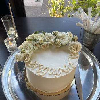 Walnut Street Kitchen - A round white cake decorated with white roses and buds on top, with the word "Vogued" written in white icing on the surface. The cake is placed on a silver tray on a dark table, with a knife beside it. In the background, there are candles, a glass with a straw, and a container holding white plastic utensils. Outside the window, greenery and a sidewalk are visible. - Salad Near Me - Montclair, New Jersey