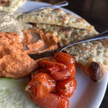 SPIN! Pizza - Roasted red pepper/ Hummus appetizer dish... - The image shows a white plate with Indian cuisine items, including naan bread topped with herbs, a serving of spicy tomato chutney, roasted cherry tomatoes, and a slice of cucumber. The plate is placed on a dark wooden table, with utensils and a napkin visible in the background. The focus is on the food, highlighting its textures and colors. - Pizza Near Me - Lawrence, Kansas
