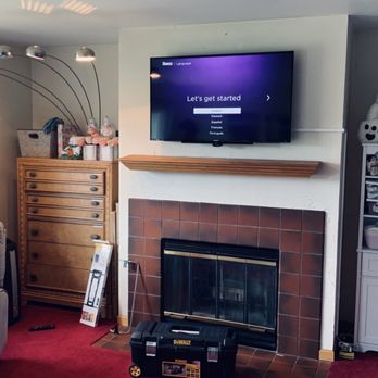 TV Mounting Same Day - A man wearing a cap and a light-colored shirt stands in a living room, holding a tool or object. The room features a fireplace with a tiled surround, a wall-mounted flat-screen TV displaying a setup screen, and a white shelving unit filled with toys, books, and storage bins. There is a window with curtains on the left, and various household items are visible, including a box and a remote control on the floor. - TV Mounting Near Me - Aurora, Illinois