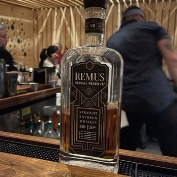 Osa - Remus vii - A bottle of Remus Repeal Reserve bourbon whiskey, labeled as straight bourbon with 50% alcohol, is positioned on a wooden bar counter. The bottle is partially filled with dark amber liquid. The background shows a bar setting with wooden decor, bar tools, and patrons, some wearing masks, and a bartender. The ceiling features wooden slats arranged in a pattern. - Cocktail Bars Near Me - Middletown, Connecticut