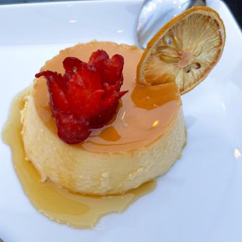 Debajo Tapas ABQ - A creamy flan dessert with caramel sauce is presented on a white plate, topped with a dried lemon slice and a red fruit garnish. A metal spoon rests nearby, and the plate is positioned near a window, suggesting a dining setting. - Spanish Near Me - Albuquerque, New Mexico
