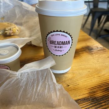Breadman Miami - Cafe Frio - A large disposable coffee cup with a lid from Breadman Miami bakery is positioned on a wooden table. The cup features a pink and black logo with the text 'Breadman Miami Bakery' and the slogan 'Eat. Love. Bread.' In the background, part of a smaller cup and a wrapped food item are visible, along with a person and some chairs, indicating a casual dining or takeaway setting. - Bakeries Near Me - Miami, Florida