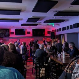 Can't beat a good live band to increase the fun quotient... - The image shows a bar with a stage where a live band is performing. Patrons are seated at tables and standing, watching the performance. The bar area has a brick wall background, multiple large TVs mounted on the walls, and a ceiling with black acoustic panels. The atmosphere appears lively with people enjoying music and drinks.