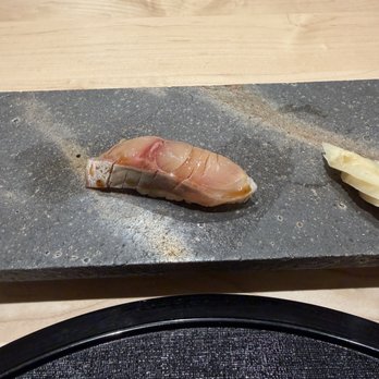 Two Nine - A piece of raw fish sushi is placed on a black stone plate, with other sushi pieces visible in the background on a wooden surface. - Sushi Bars Near Me - Washington, DC, District of Columbia