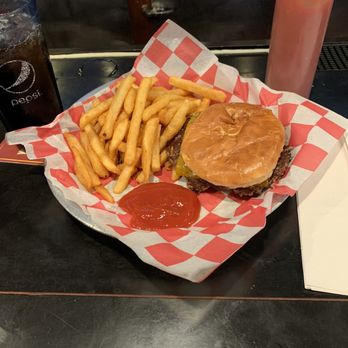 Echo Tap & Grill - My Dinner. Burger and fries. - A burger with a toasted bun, pickles, and a beef patty is served on a tray lined with red and white checkered paper. Accompanying the burger are French fries and a small portion of ketchup. A glass of soda and a squeeze bottle of ketchup are also visible on the dark table surface. The setting appears to be a casual dining restaurant. - Sports Bars Near Me - Madison, Wisconsin
