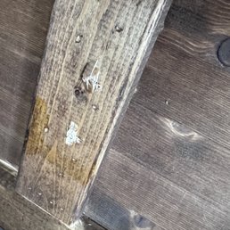 wood screws attaching the table top were ripped loose. - The image depicts a close-up of a wooden surface, likely part of furniture or flooring, with a frame or border. The wood shows signs of damage, including ripped loose screws and some peeling or chipped areas. The surface has a natural wood grain pattern and appears aged or worn.