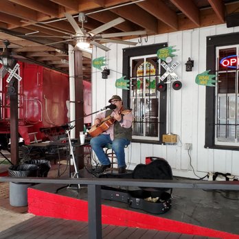The Loose Caboose - A man wearing sunglasses and a cap plays an acoustic guitar and sings into a microphone on a small stage with a white wall and window behind him. - Barbeque Near Me - Spring, Texas