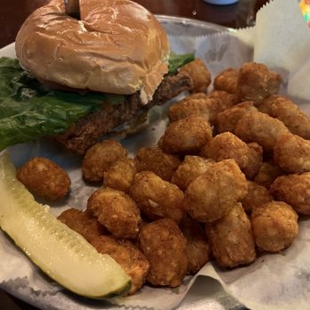 Brazen Head Pub - Pork schnitzel Tuesday special with cracked pepper tater tots. - A meal featuring a burger with lettuce and a bun is accompanied by a large portion of fried tater tots and a pickle spear. The food is presented on a tray lined with parchment paper, with a dark wooden table visible in the background. - Pubs Near Me - West Bend, Wisconsin