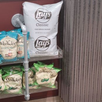 Cousins Subs - Classic Lay's chips - A large bag of Lay's Classic potato chips is positioned on a metal shelf in a store setting. The bag is white with black and gray branding. Smaller bags of Miss Vickie's Jalapeño chips are also visible on the shelf below. The shelf is attached to a wall with a pipe running vertically beside it, and the background includes a textured wall panel and a countertop. - Sandwiches Near Me - Monona, Wisconsin