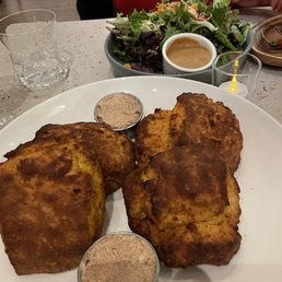 Sweet potato bread - A white plate holds three fried chicken pieces with a golden-brown, crispy exterior. Two small metal bowls contain a seasoning or dipping sauce. In the background, there is a bowl of salad with mixed greens and a small container of dressing, along with a glass of water and a glass of a dark beverage. The setting appears to be a restaurant with a light-colored table surface.