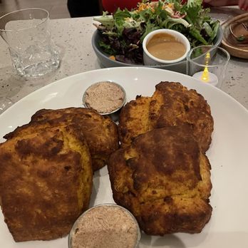 BLDG - Sweet potato bread - A white plate holds three fried chicken pieces with a golden-brown, crispy exterior. Two small metal bowls contain a seasoning or dipping sauce. In the background, there is a bowl of salad with mixed greens and a small container of dressing, along with a glass of water and a glass of a dark beverage. The setting appears to be a restaurant with a light-colored table surface. - New American Near Me - New Haven, Connecticut