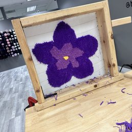 A purple flower-shaped tufted rug is mounted on a wooden frame at a DIY rug tufting studio, with purple yarn scraps on the table.
