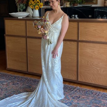 Loulette Bride - A woman wearing a white satin wedding dress with a train stands indoors in front of a wooden counter. She holds a bouquet of dried flowers and has dark hair styled in an updo. The background features a wooden counter, a large floral arrangement with sunflowers, and framed artwork on the wall. The setting appears to be a boutique or event space with warm lighting and decorative elements. - Bridal Near Me - Brooklyn, New York