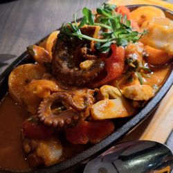 Gordion - "Fisch Sote" in der gusseisernen Pfanne mit Lachs und Weißfisch sowie Tintenfisch und zwei Scampis auf Spitzpaprika, Knoblauch und Zwiebeln