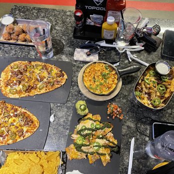 Topgolf - brisket flatbread, buffalo chicken dip, chicken nachos, macaroni bites, cheese fries - Food items such as two pizzas, nachos, fried appetizers, and chips are displayed on a table at Topgolf, with drinks and condiments nearby. - Golf Near Me - Ashburn, Virginia
