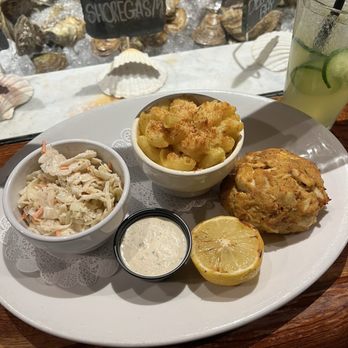 Mama's on the Half Shell - A seafood platter features macaroni and cheese, coleslaw, a lemon half, and a creamy dipping sauce, with a drink containing cucumber slices in the background. - Bars Near Me - Baltimore, Maryland