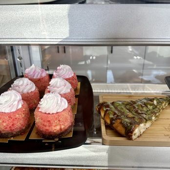 Mocha Me - The image features a glass display case containing baked goods. On the left, there are six pink cupcakes with white frosting, arranged on a black tray. To the right, there is a slice of dessert with a chocolate drizzle and green topping, placed on a wooden board. The background shows reflections of the outside environment through the glass. - Coffee & Tea Near Me - Melvindale, Michigan