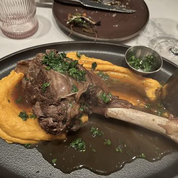 Brooklyn Winery - Lamb shank - Lamb shank with gravy is presented on a black plate, accompanied by mashed potatoes and garnished with herbs, in a restaurant setting. - Venues & Event Spaces Near Me - Brooklyn, New York