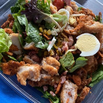 Chick-fil-A - Cobb Salad - A Cobb salad in a clear plastic container contains fried chicken pieces, cherry tomatoes, sliced hard-boiled eggs, bacon bits, shredded cheese, corn, and mixed greens. The salad is assembled with various fresh vegetables and protein ingredients, typical of a Cobb salad served as a meal option. - Fast Food Near Me - Wyncote, Pennsylvania