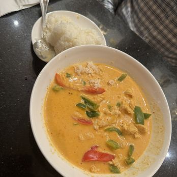 Maya Thai Eatery - Panang Curry chicken - A white bowl contains Thai curry with a creamy, orange-colored sauce, including vegetables like tomatoes and green peppers, and pieces of chicken. A small dish with white rice and a spoon is placed nearby. The setting appears to be a restaurant with a dark, speckled table surface and a person wearing a plaid shirt seated nearby. - Thai Near Me - Westfield, New Jersey
