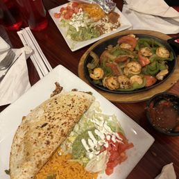 Grilled Chicken Quesadillas Fajitas, Shrimp Fajitas - The image shows a Mexican meal with a quesadilla, shrimp fajitas, and a salad on a wooden table, with drinks and utensils.
