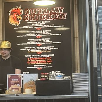 Outlaw Chicken - A black menu board for Outlaw Chicken lists menu items including Nashville hot chicken, half and quarter rotisserie chicken, chicken sandwiches, and wings with prices. The board features a stylized rooster logo and is illuminated by a hanging light. There are additional items like sides and sauces at the bottom, with a person standing nearby and some condiments on the counter. - Chicken Shop Near Me - Albuquerque, New Mexico