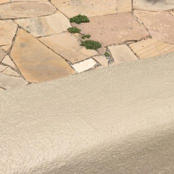 Always Construction Services - a closeup of a sidewalk - The photo captures a beige textured surface, possibly a wall or a ledge, next to a stone-paved area composed of irregularly shaped stones. Small green plants are growing in the cracks between the stones, indicating outdoor conditions and some plant growth in the area. - Stucco Services Near Me - Santa Fe, New Mexico
