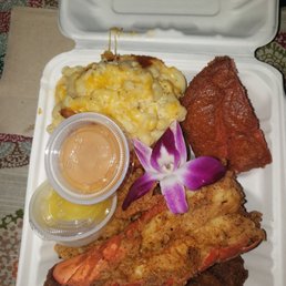 Fried Shrimp - A seafood platter in a white takeout box includes fried shrimp, macaroni and cheese, a piece of fried fish, and a flower garnish, with small containers of sauce.