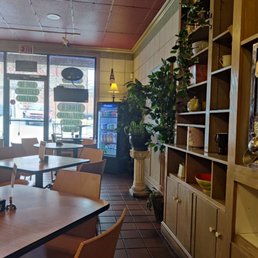 Nice atmosphere - The image shows the interior of a Mediterranean restaurant with a tiled floor and a ceiling with acoustic tiles. Wooden shelves with decorative items and plants line the right wall. Near the entrance, there is a vending machine stocked with beverages, and a small lamp on a pedestal. Several tables with chairs are arranged for diners, and large windows at the front allow natural light to enter. The restaurant has a cozy, decorated atmosphere with Mediterranean-themed decor.