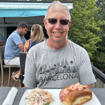 Patio 03907 - Lobster roll with cole slaw - A man with sunglasses and a gray Barcelona t-shirt is sitting outdoors at a table with two plates of food, smiling at the camera. In the background, other people are seated at a table under a canopy, with trees surrounding the area. - Seafood Near Me - Ogunquit, Maine