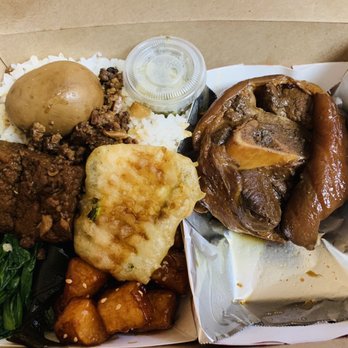 AhChung’s Kitchen - Pork leg set meal - A Chinese-style takeout box holds rice, braised pork, a boiled egg, fried tofu, greens, and other dishes, with a small container of sauce. - Chinese Near Me - Aurora, Illinois