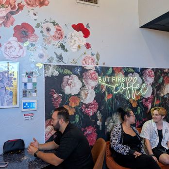 Inspired Brew - Inside - The image shows three individuals seated inside a coffee shop. One person is sitting at a small round table, facing left, with a black shirt and glasses. Two others are sitting on a sofa against a floral-patterned wall, engaged in conversation. The wall features large floral artwork and a neon sign that says 'But First Coffee.' There is a security camera and a small display on the wall, indicating a modern, stylish interior. - Coffee & Tea Near Me - Dallas, Texas