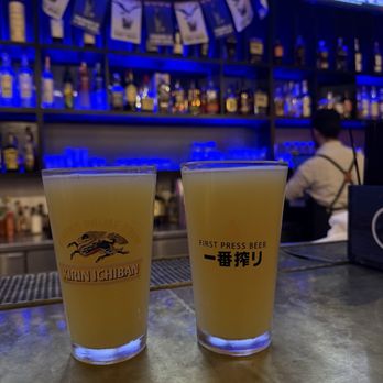 Behind Bars - Two beers! - Two glasses of beer sit on a bar counter, with a bar and television screens displaying sports in the background. The bar has bottles and a bartender. - Bars Near Me - Pasadena, California