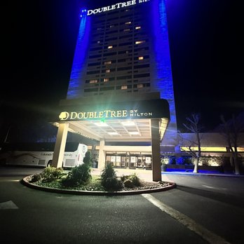 DoubleTree by Hilton Hotel Albuquerque - Road trip 4th stop - Albuquerque NM - The image shows a multi-story DoubleTree by Hilton hotel at night, with the building illuminated by blue lighting along its sides. The hotel sign is prominently displayed above the entrance, which is lit and features the DoubleTree logo. The foreground includes a small landscaped area with plants and a parking lot, with a bus visible on the left side. The overall scene emphasizes the hotel's exterior lighting and signage. - Hotels Near Me - Albuquerque, New Mexico