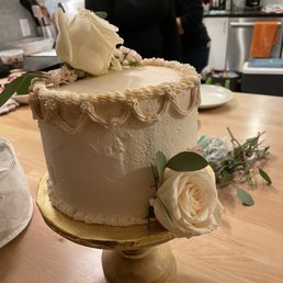 Cake! - A white, textured-frosted cake decorated with cream-colored roses and green leaves is displayed on a gold cake stand. The cake features piped icing details along the top and bottom edges. It is placed on a wooden table in a kitchen environment, with people and kitchen appliances visible in the background.