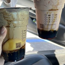Rose Milk Tea - Two bubble tea drinks with toppings are held by a person inside a vehicle. The drinks are in clear plastic cups with measurement markings and QR codes, containing dark tapioca pearls at the bottom, with one drink having a yellow pudding topping and the other a pinkish cream topping. The background shows part of the car interior and sunlight coming through the window.