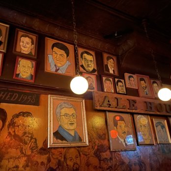 Old Town Ale House - The image shows a wall inside a bar decorated with numerous framed portraits of various individuals, some of which are caricatures or stylized illustrations. The portraits are arranged in a somewhat haphazard manner, with some hanging from the ceiling by chains and others mounted directly on the wall. The setting appears to be a bar or pub with a dark, wooden interior, and there are two spherical light fixtures hanging from the ceiling. The wall features a mural or painting with multiple faces, and a wooden sign with the word 'ALEHOUSE' is partially visible. The overall atmosphere suggests a vintage or eclectic decor style. - Pubs Near Me - Chicago, Illinois