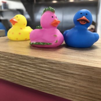 Duck Donuts - My fave hawked duck - Three rubber duck figurines in pink, yellow, and blue are arranged on a wooden surface, with a kitchen background. - Coffee & Tea Near Me - Orange, Connecticut