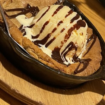 Chili's - Skillet cookie - A dessert featuring a large, baked skillet cookie with a golden-brown crust, topped with a scoop of vanilla ice cream. The ice cream is drizzled with chocolate syrup, and additional chocolate syrup is spread over the cookie. The skillet is placed on a round wooden tray, and the background includes a wooden table surface. - Bars Near Me - Raynham, Massachusetts