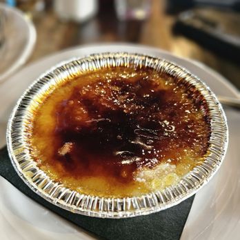 Range Cafe - Creme brulee - A crème brûlée dessert with a caramelized sugar crust is presented in a small aluminum container. The dish is placed on a white plate with a black napkin underneath. The background shows a blurred restaurant setting with various objects and condiments. - Breakfast & Brunch Near Me - Albuquerque, New Mexico