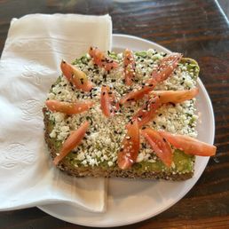 Avocado Toast - A piece of avocado toast on a white plate, garnished with cherry tomato slices, crumbled cheese, and black sesame seeds, served with a napkin.
